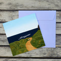 A square art print or postcard showing a coastal landscape with green hills, a winding tan path, dark blue water, and a pale blue sky, placed over a light lavender envelope on a weathered wood surface.